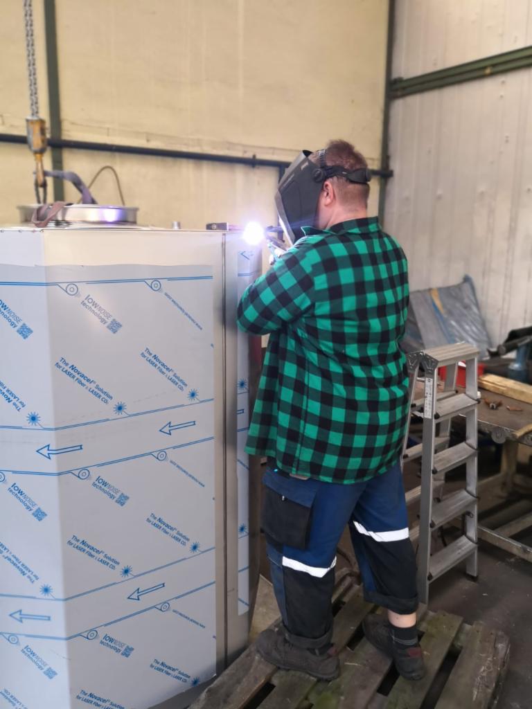 Pracownik w masce spawalniczej i zielonej koszuli w kratę spawa metalową obudowę, widoczne iskry i odbicia światła na powierzchni metalu.