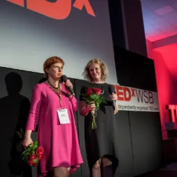 Dwie kobiety na scenie podczas wydarzenia TEDxWSB, jedna w różowej sukience z mikrofonem, druga w czarnej sukience z bukietem czerwonych róż.