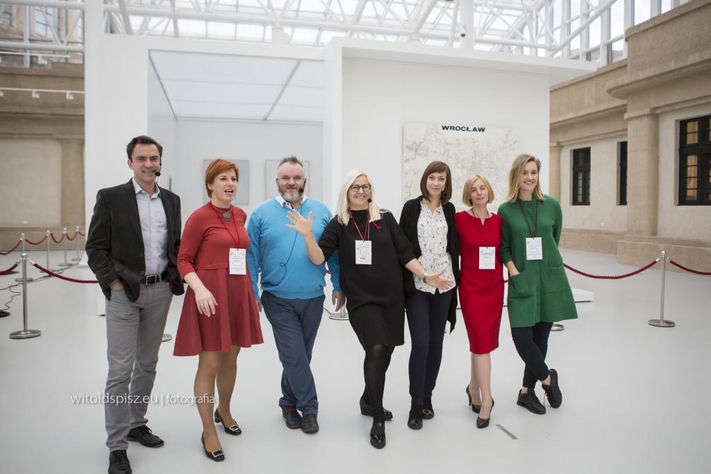 Grupa osób, prawdopodobnie uczestników lub prelegentów, pozuje na tle mapy Wrocławia w jasnym, nowoczesnym wnętrzu. Część osób ma mikrofony, a wszyscy mają identyfikatory na szyjach.