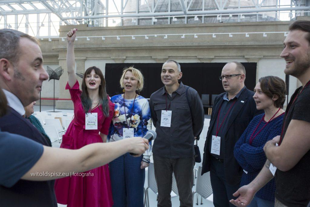 Grupa osób w trakcie interaktywnego szkolenia, jedna z kobiet w różowej sukience z podniesioną ręką, mikrofon w kadrze, identyfikatory na szyjach uczestników.