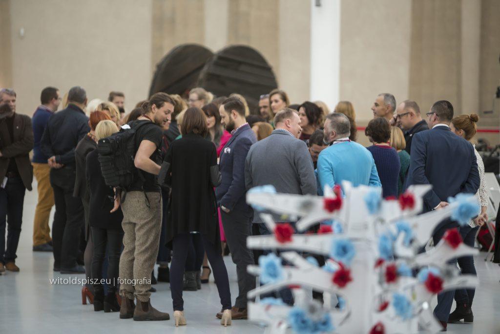 Grupa osób uczestnicząca w wydarzeniu, być może konferencji lub szkoleniu, z dekoracjami kwiatowymi w pierwszym planie i artystyczną instalacją w tle.