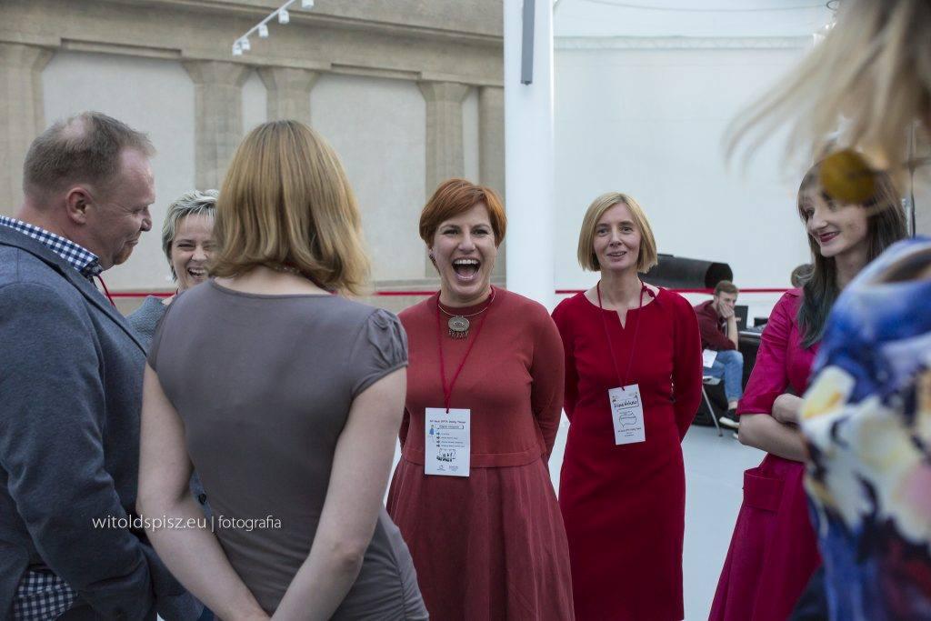 Grupa osób w trakcie interakcji podczas wydarzenia, kobiety w czerwonych sukienkach z identyfikatorami, mężczyzna w marynarce, widoczne uśmiechy i zaangażowanie.