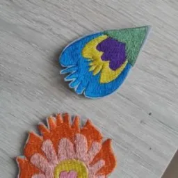 Dwie naszywki: pióro pawie z niebieskim, żółtym i fioletowym haftem oraz kwiat w odcieniach pomarańczy i różu, leżące na jasnoszarym blacie.