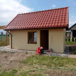Niewielki budynek gospodarczy z nową, czerwoną dachówką i beżową elewacją, widoczny z perspektywy trawnika, z czerwonym krzesłem i narzędziami obok wejścia.