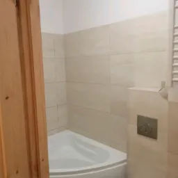 Wykończona łazienka z narożną wanną, beżowymi płytkami imitującymi kamień i drewnianą ościeżnicą drzwiową z widocznymi sękami.