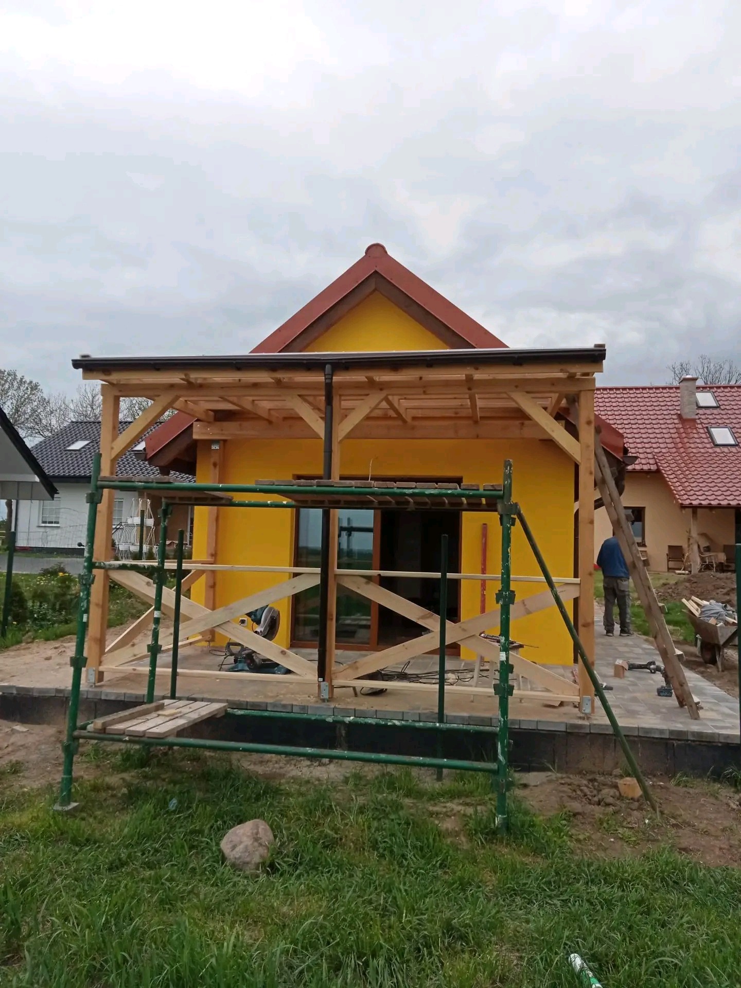 Żółty dom z brązowym dachem w trakcie budowy z drewnianą konstrukcją werandy, widoczne rusztowania i pracownik z drabiną w tle.