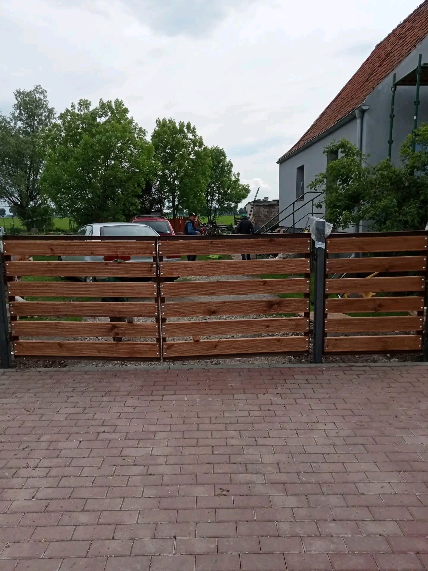 Nowoczesne, drewniane ogrodzenie panelowe z metalowymi słupkami na tle domu z czerwoną dachówką, widoczne samochody i osoby w tle.