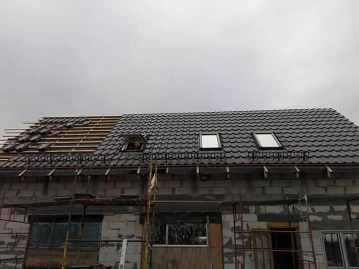 Budowa domu w trakcie: widoczna część dachu pokryta ciemną dachówką, okna dachowe zamontowane, druga część dachu z widoczną konstrukcją drewnianą, ściany z pustaków częściowo otynkowane, rusztowania.