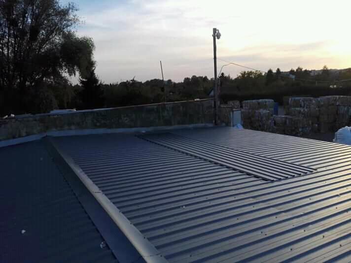 Widok z góry na nowy, blaszany dach z widocznymi łączeniami i elementami montażowymi, na tle drzew i zachodzącego słońca.