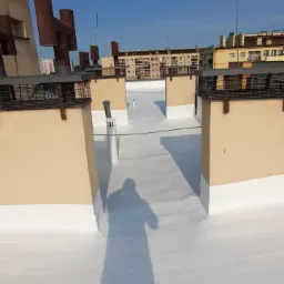 Biała, hydroizolacyjna powłoka na dachu budynku z widocznymi kominami i antenami. W tle zabudowa miejska i błękitne niebo. Cień osoby na pierwszym planie.