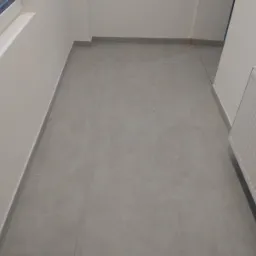 Szare, kwadratowe płytki podłogowe ułożone w jasnym korytarzu z grzejnikiem i listwami przypodłogowymi. Widoczny fragment okna. Minimalistyczny wystrój.