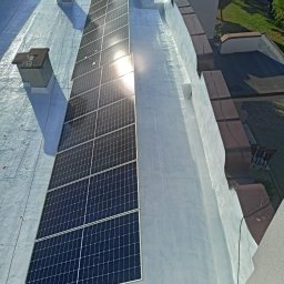 F.U.Baks-Bud - Biały dach budynku z panelami słonecznymi w słoneczny dzień. Widok z góry ukazuje kominy i elementy architektoniczne. Część paneli odbija światło.