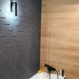 Nowoczesna łazienka z dwiema kontrastującymi ścianami: szara ściana z trójwymiarowymi płytkami i lampą ścienną oraz ściana wyłożona płytkami imitującymi jasne drewno, widoczny fragment kabiny...