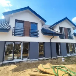 Dwukondygnacyjny dom z elewacją w kolorze kremowym i szarym, z drewnianymi panelami dekoracyjnymi na piętrze, czarną dachówką i elementami wykończeniowymi, widok z zewnątrz na plac budowy...