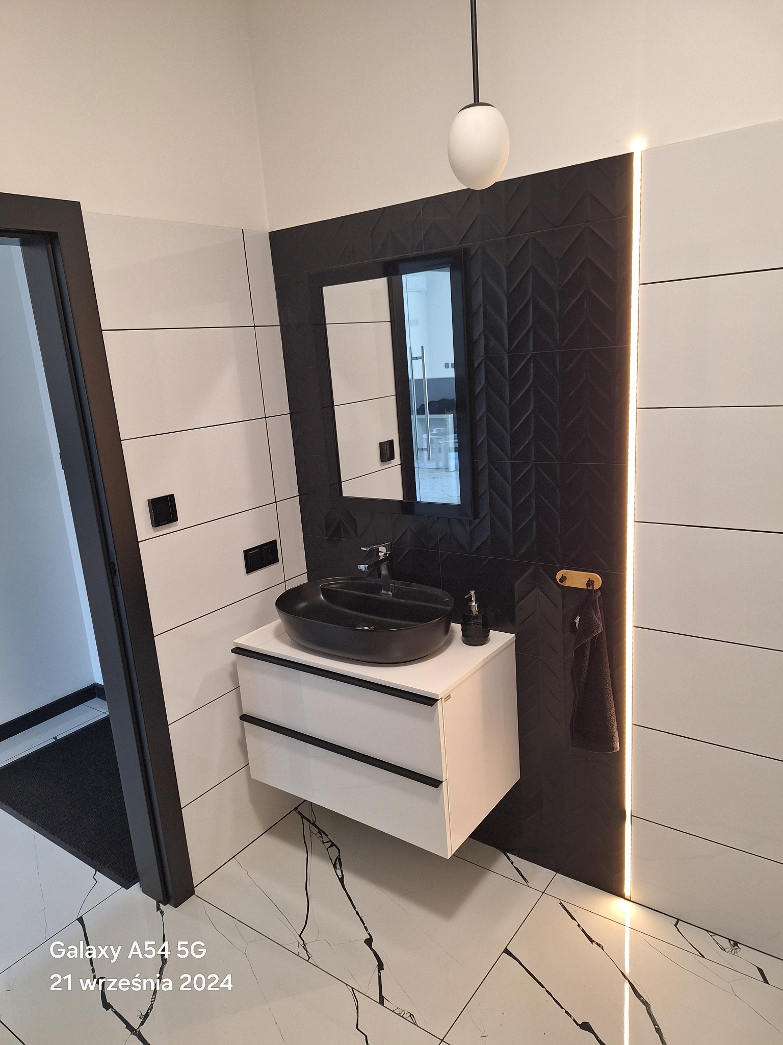 Nowoczesna łazienka z czarną umywalką nablatową, białymi płytkami ściennymi i podłogą imitującą marmur z czarnymi żyłami; dekoracyjna ściana z czarnych płytek 3D w jodełkę i podświetleniem LED.