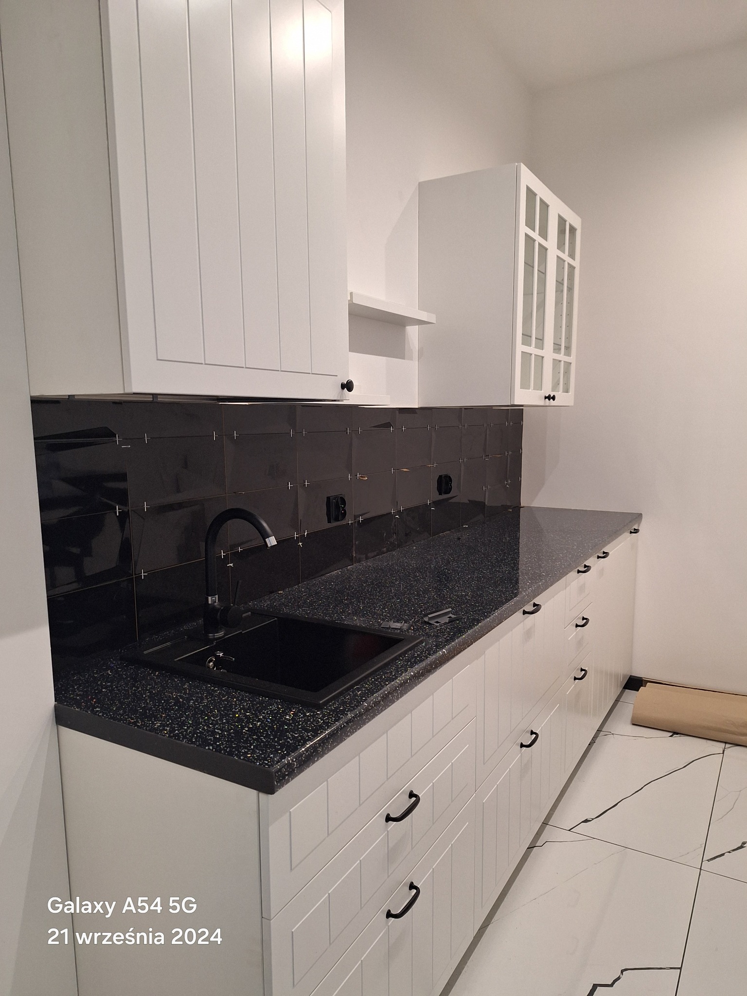 Nowoczesna kuchnia z czarnym blatem i błyszczącymi czarnymi płytkami nad zlewem, białe szafki z frezowanymi frontami i czarnymi uchwytami, marmurowa podłoga.