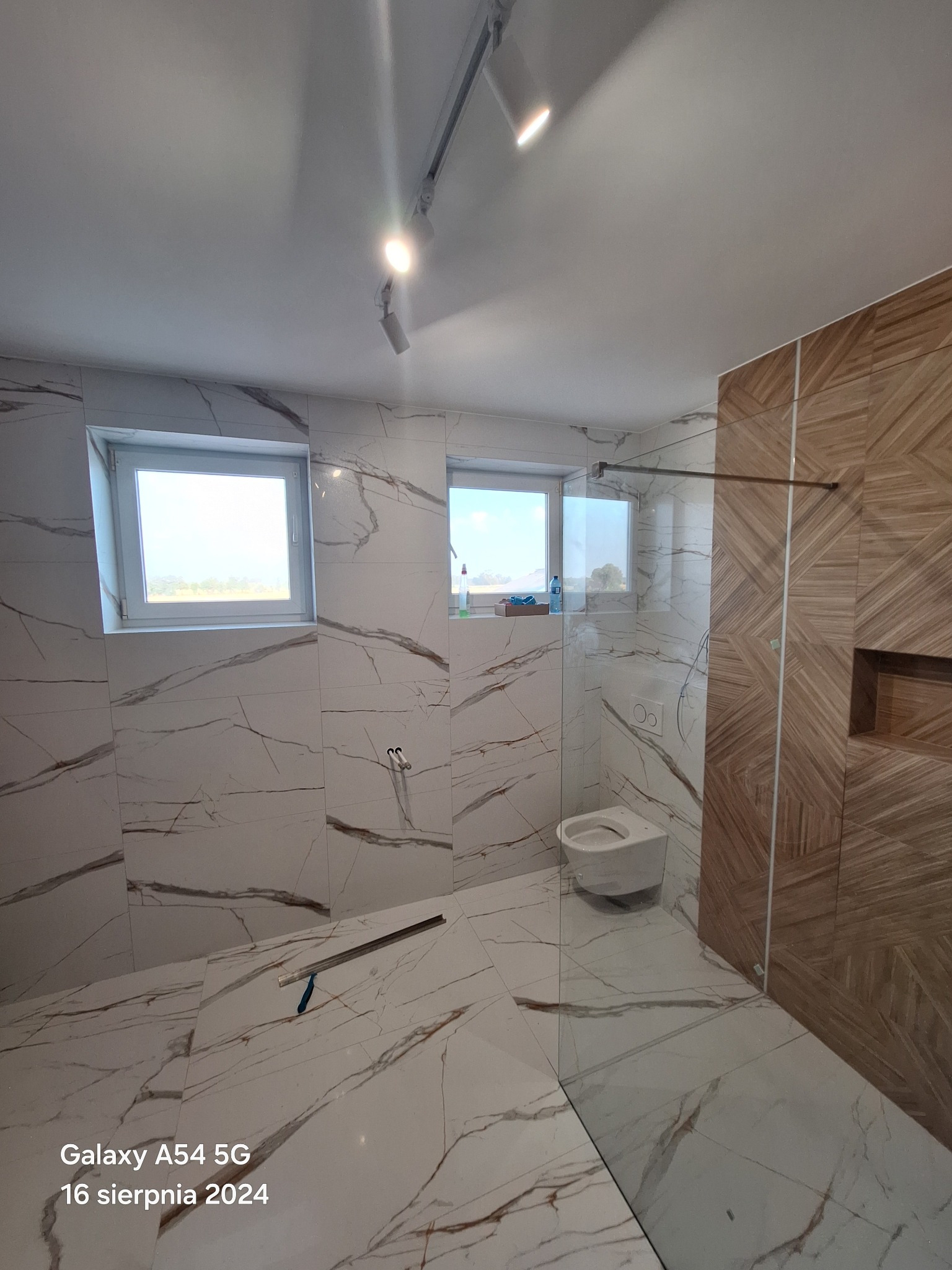 Nowoczesna łazienka z marmurowymi płytkami, szklaną kabiną prysznicową i drewnianą ścianą akcentującą, widoczne narzędzia na podłodze sugerują trwające prace wykończeniowe.