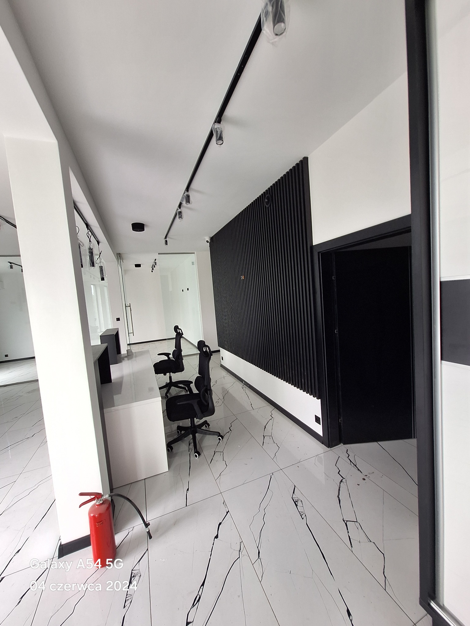 Nowoczesne biuro z białymi ścianami, marmurową podłogą, czarnymi akcentami w postaci lameli na ścianie i oświetlenia szynowego, wyposażone w biurka i krzesła biurowe, z widoczną gaśnicą w rogu.