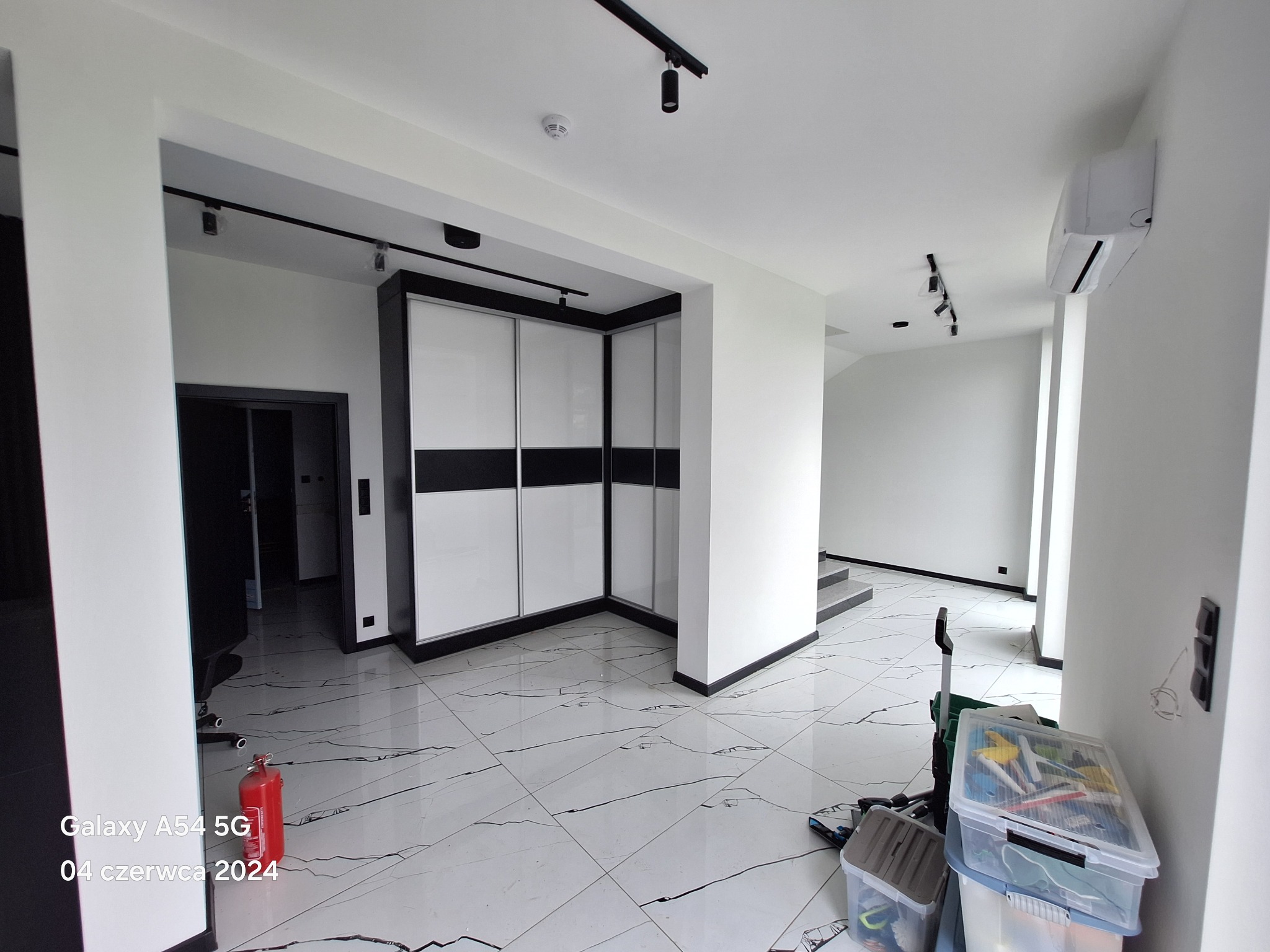 Wnętrze przestronnego, nowocześnie wykończonego pokoju z białymi ścianami, marmurową podłogą, zabudowaną szafą z przesuwnymi drzwiami i czarnymi akcentami, widoczne narzędzia i gaśnica w rogu.