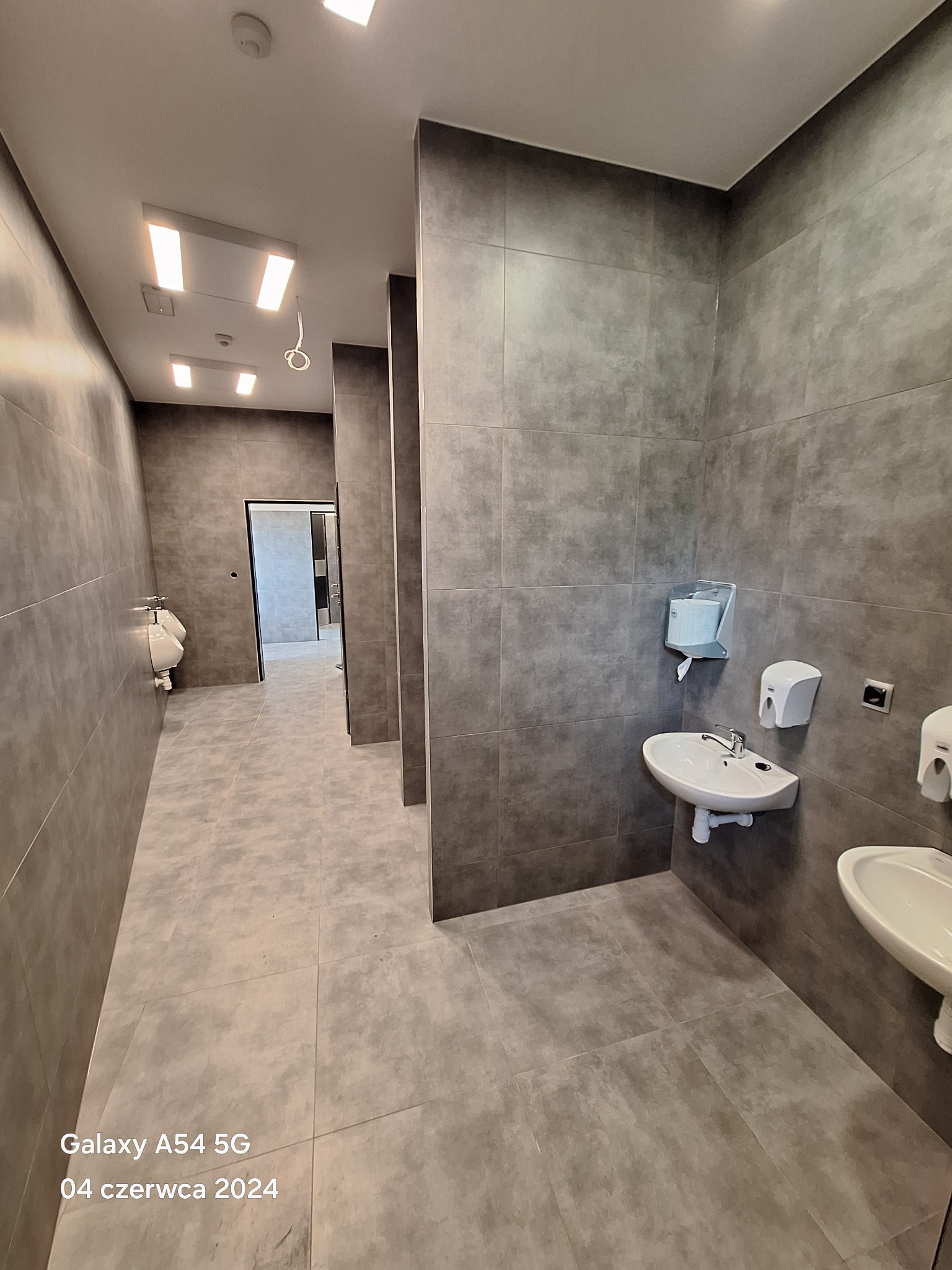 Wnętrze publicznej toalety w Głogininie z szarymi płytkami na ścianach i podłodze, widoczne umywalki z dozownikami mydła oraz pisuary w tle.