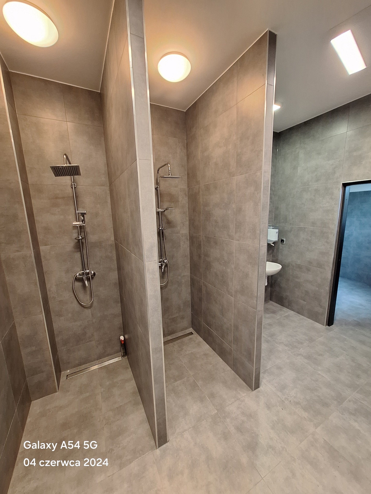 Nowoczesna łazienka publiczna z kabinami prysznicowymi wykończonymi szarą płytką, widoczny odpływ liniowy i umywalka w tle.