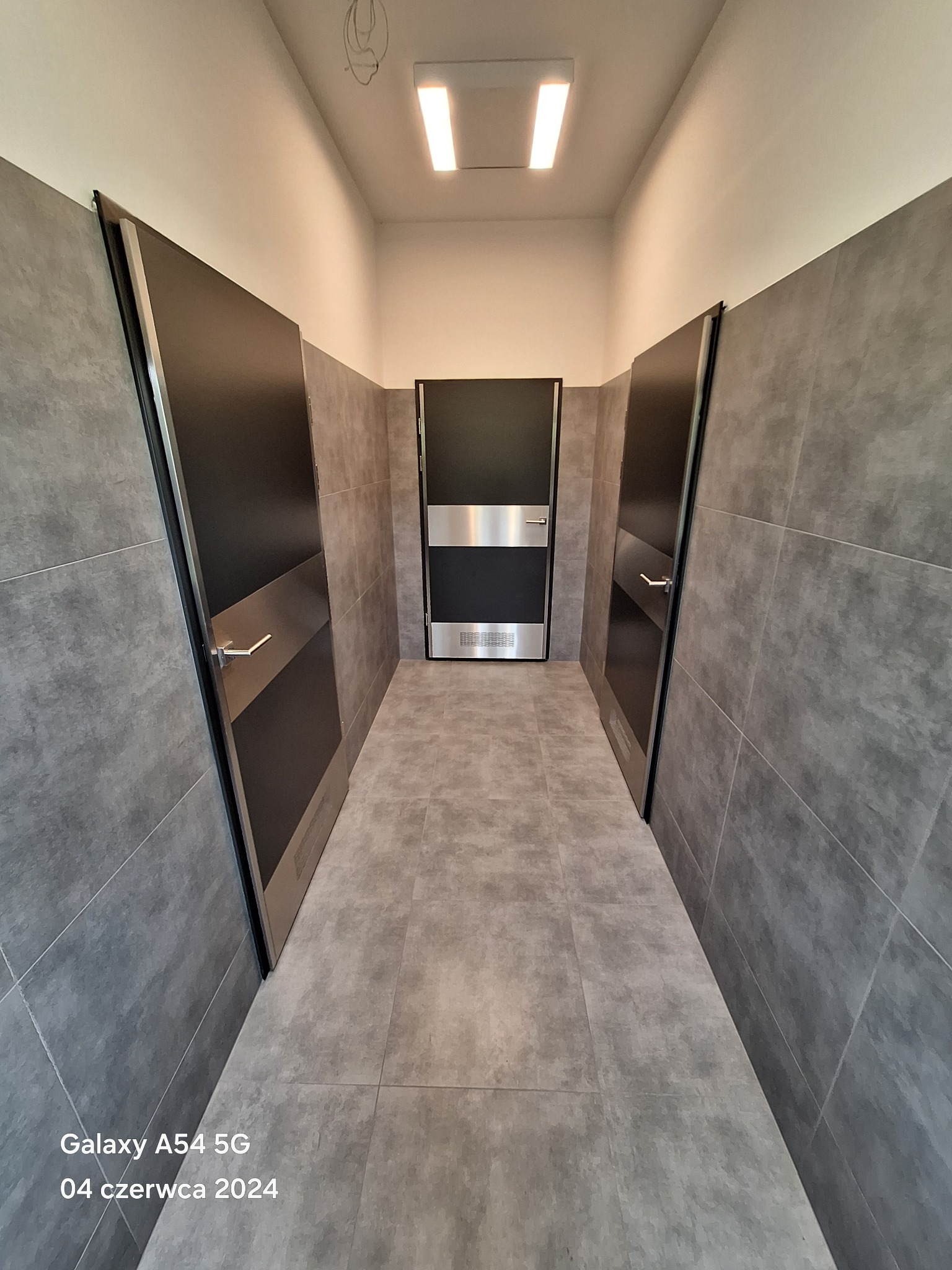 Korytarz wykończony szarymi płytkami z trzema czarnymi drzwiami z poziomymi, metalowymi akcentami, oświetlony dwoma podłużnymi lampami sufitowymi.