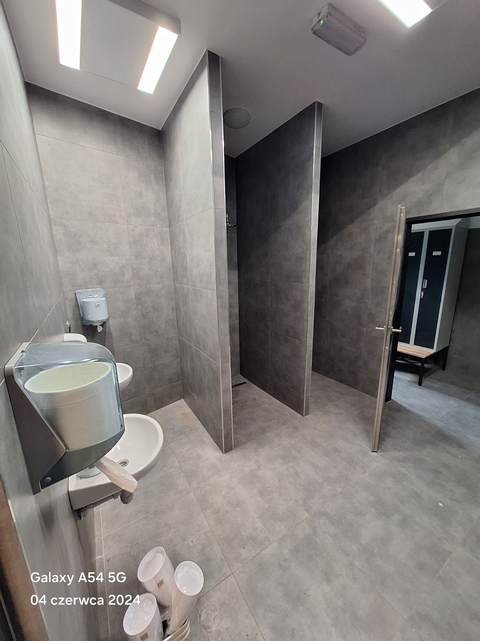 Szara łazienka publiczna z umywalkami, dozownikiem mydła, kabinami prysznicowymi i szafkami w tle.