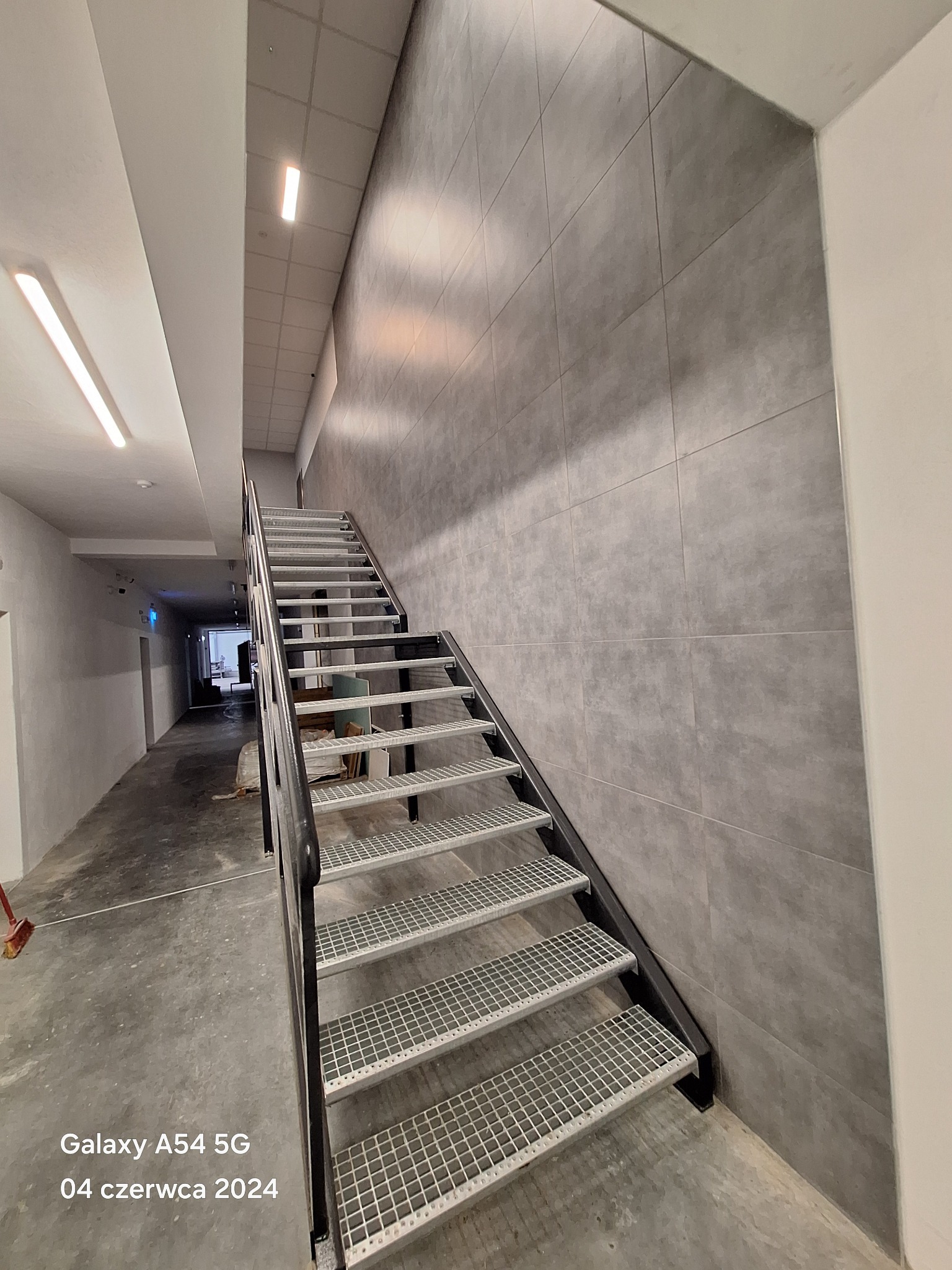 Wnętrze budynku z metalowymi schodami o ażurowych stopniach, prowadzącymi w górę wzdłuż ściany wyłożonej szarymi, prostokątnymi płytkami, widok z dołu korytarza.