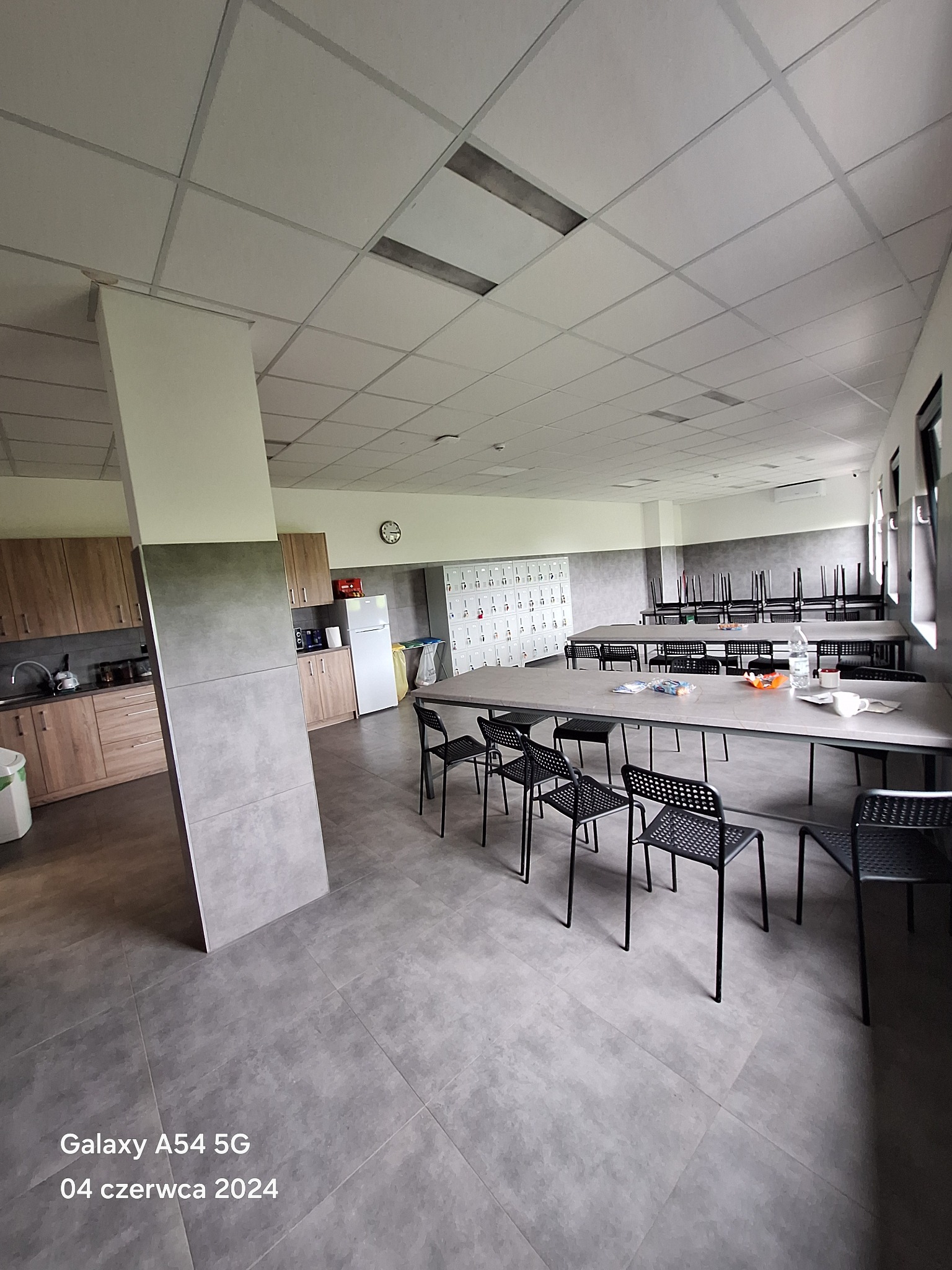 Przestronna sala z szarymi płytkami podłogowymi, stołami z metalowymi nogami i czarnymi krzesłami, widok na aneks kuchenny z drewnianymi szafkami i białą lodówką oraz ścianę z szafkami ubraniowymi.