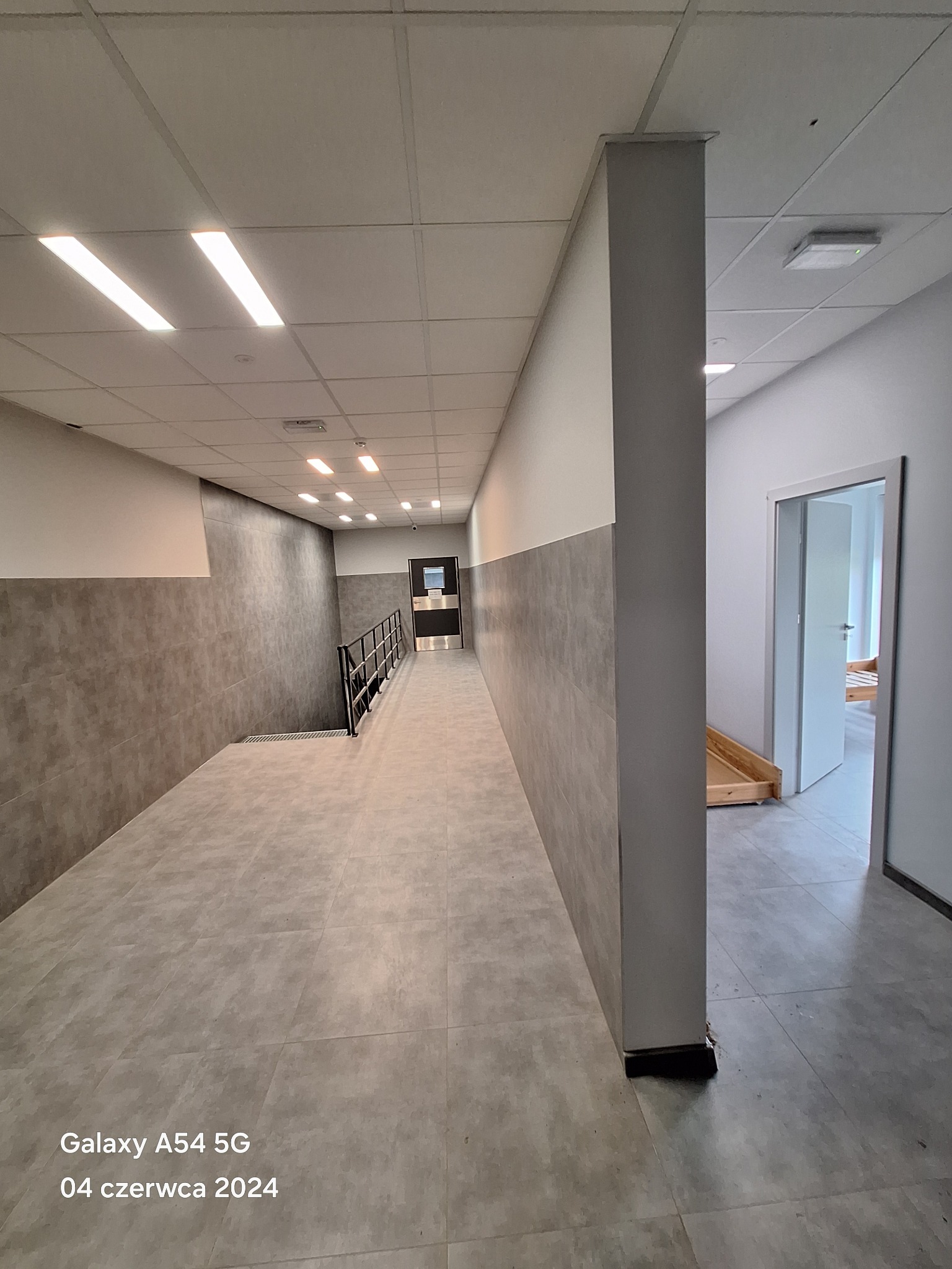 Korytarz w budynku biurowym z szarymi płytkami na podłodze i ścianach do połowy wysokości, oświetlony lampami sufitowymi.