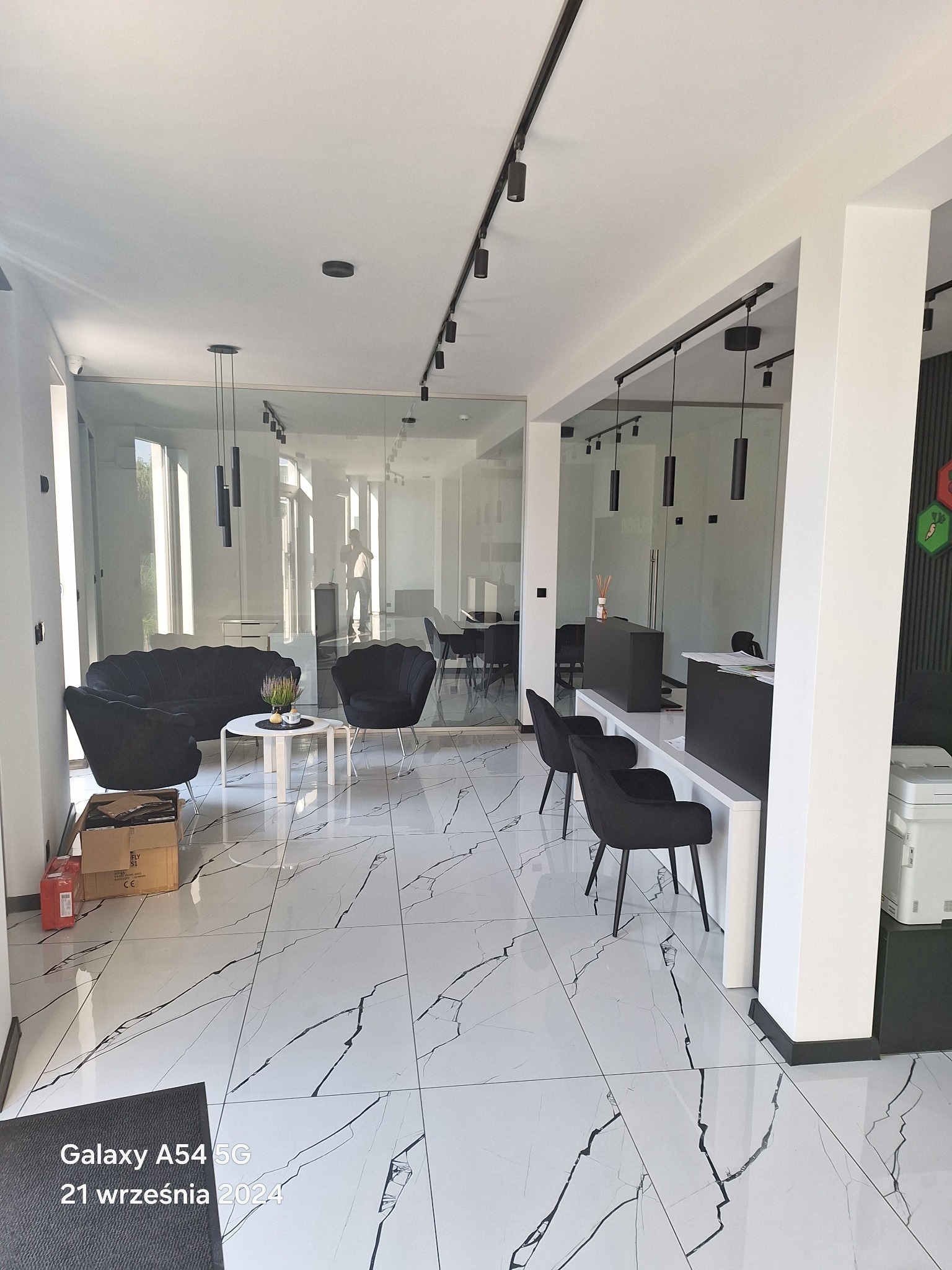 Nowoczesne wnętrze biurowe z białymi ścianami, marmurową podłogą, czarnymi meblami i szklaną ścianą działową, oświetlone reflektorami na szynoprzewodach.