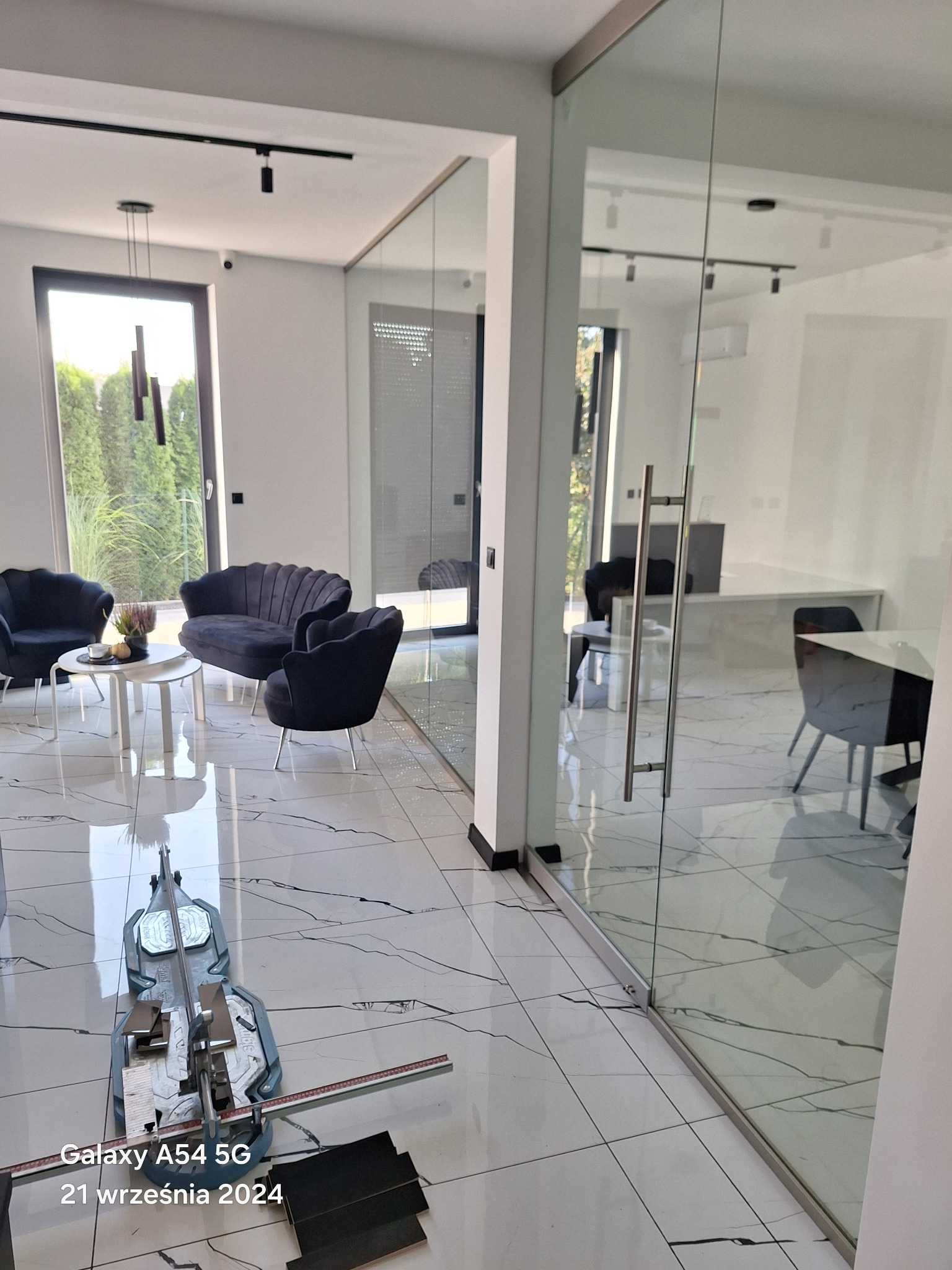 Układanie płytek podłogowych z użyciem przecinarki do płytek w nowoczesnym salonie z marmurową podłogą i szklaną ścianą działową.