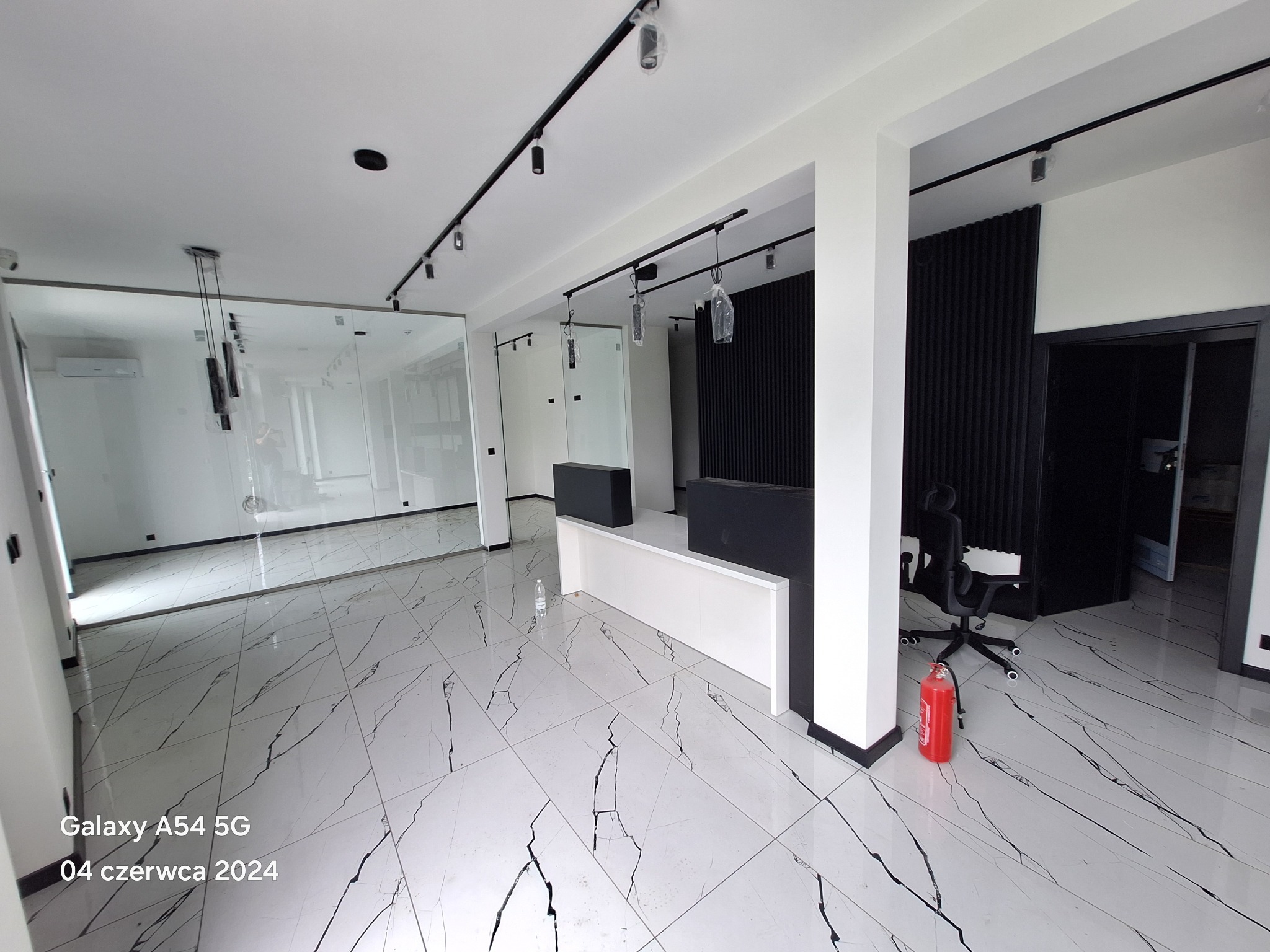 Nowoczesne wnętrze biurowe z błyszczącą białą podłogą imitującą marmur, czarnymi akcentami w postaci paneli ściennych i oświetlenia szynowego oraz szklaną ścianą działową. W tle widoczny klimatyzator.