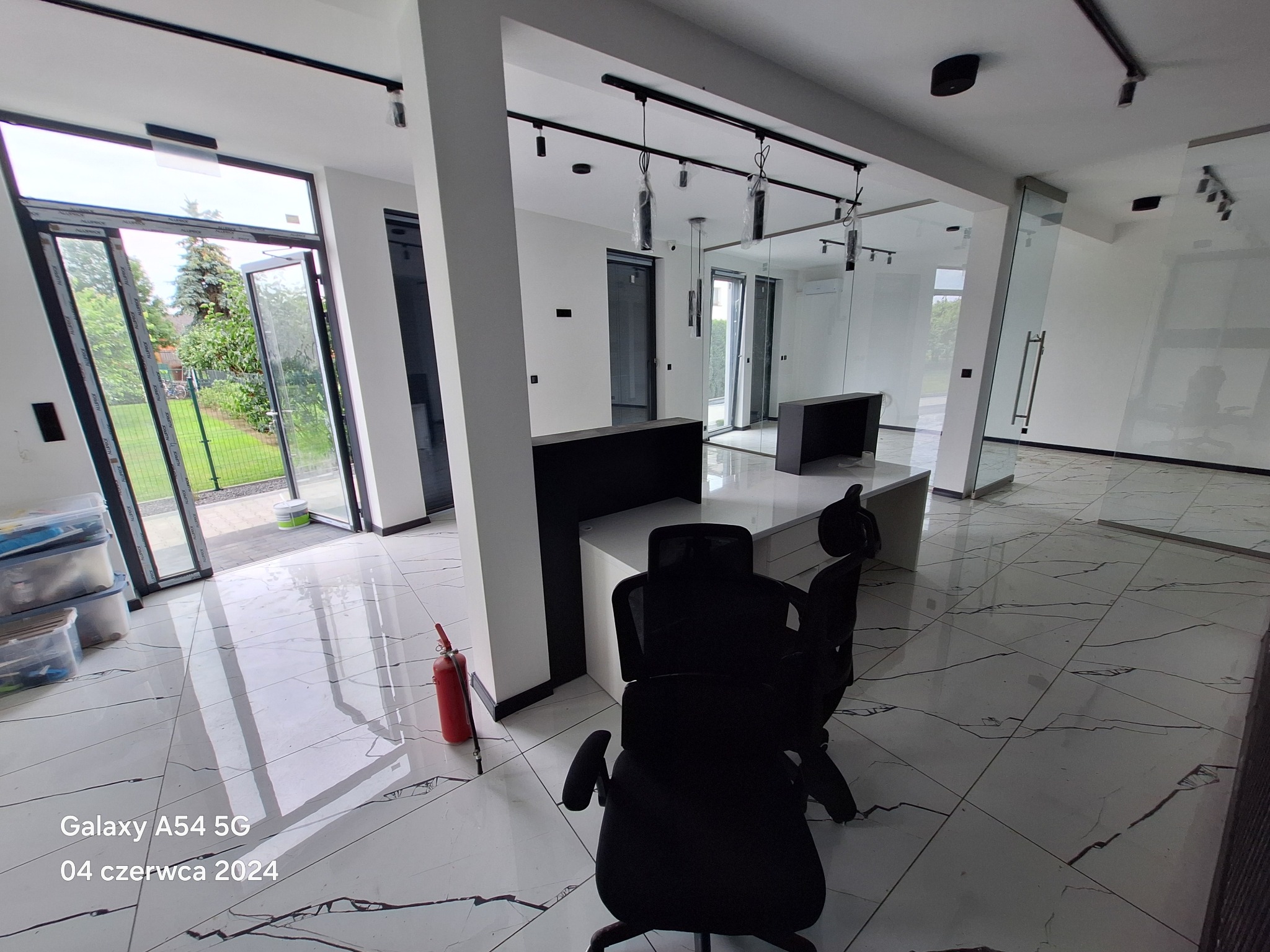 Wnętrze nowoczesnego, przestronnego biura z białymi ścianami, marmurową podłogą i czarnymi meblami, widoczne otwarte drzwi na zewnątrz i gaśnica przy kolumnie.