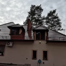 Dom z nowym, błyszczącym, brązowym dachem z dachówki ceramicznej, widoczne okna dachowe i komin z anteną na tle pochmurnego nieba i wysokich sosen.