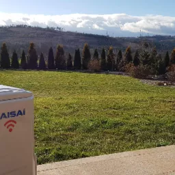 Zewnętrzna jednostka klimatyzacji marki Kaisai na tle zadbanego trawnika, ozdobnych drzew i górskiego krajobrazu pod błękitnym niebem.