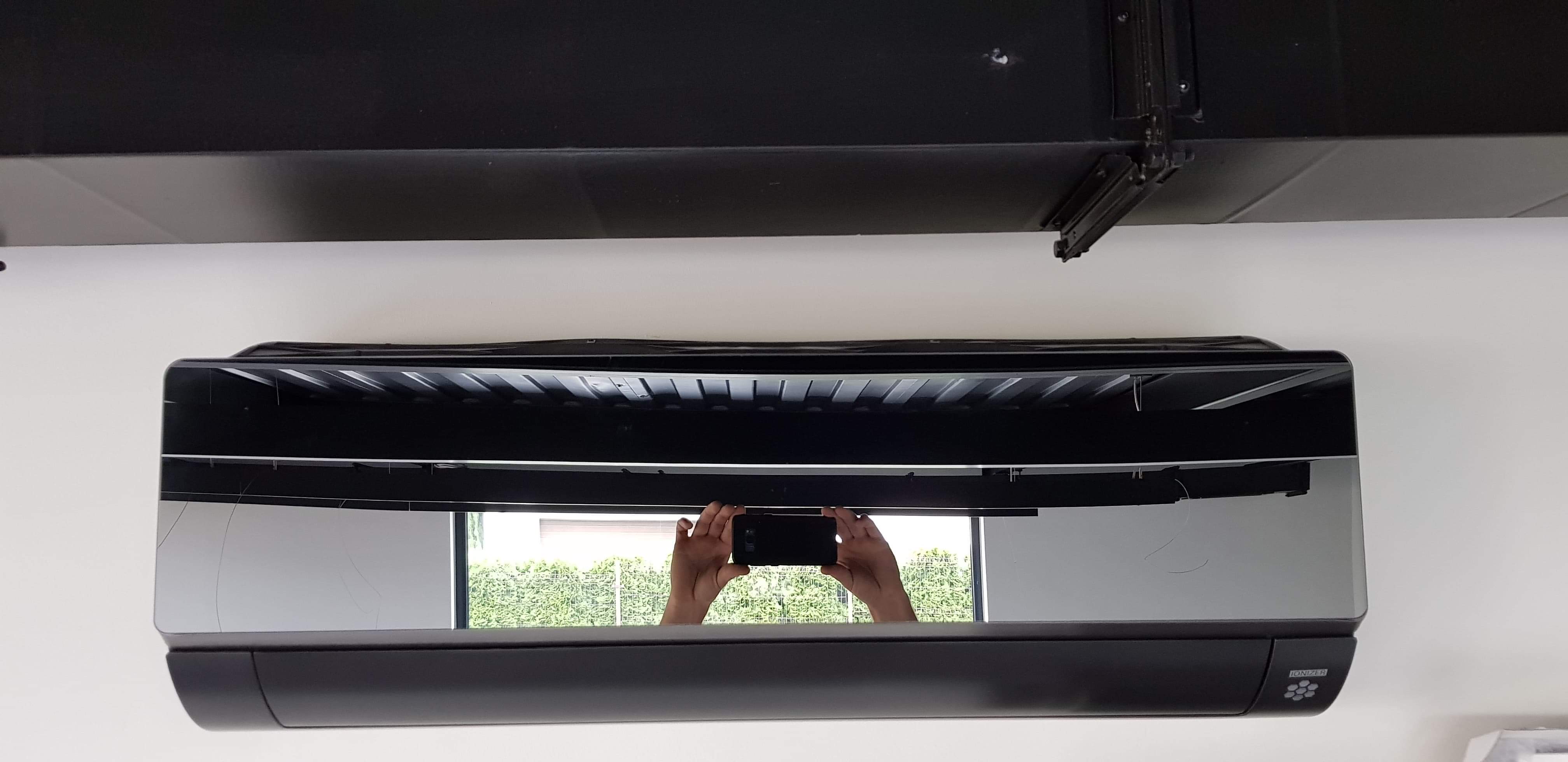 Czarny klimatyzator zamontowany pod sufitem, widoczne odbicie osoby robiącej zdjęcie telefonem.