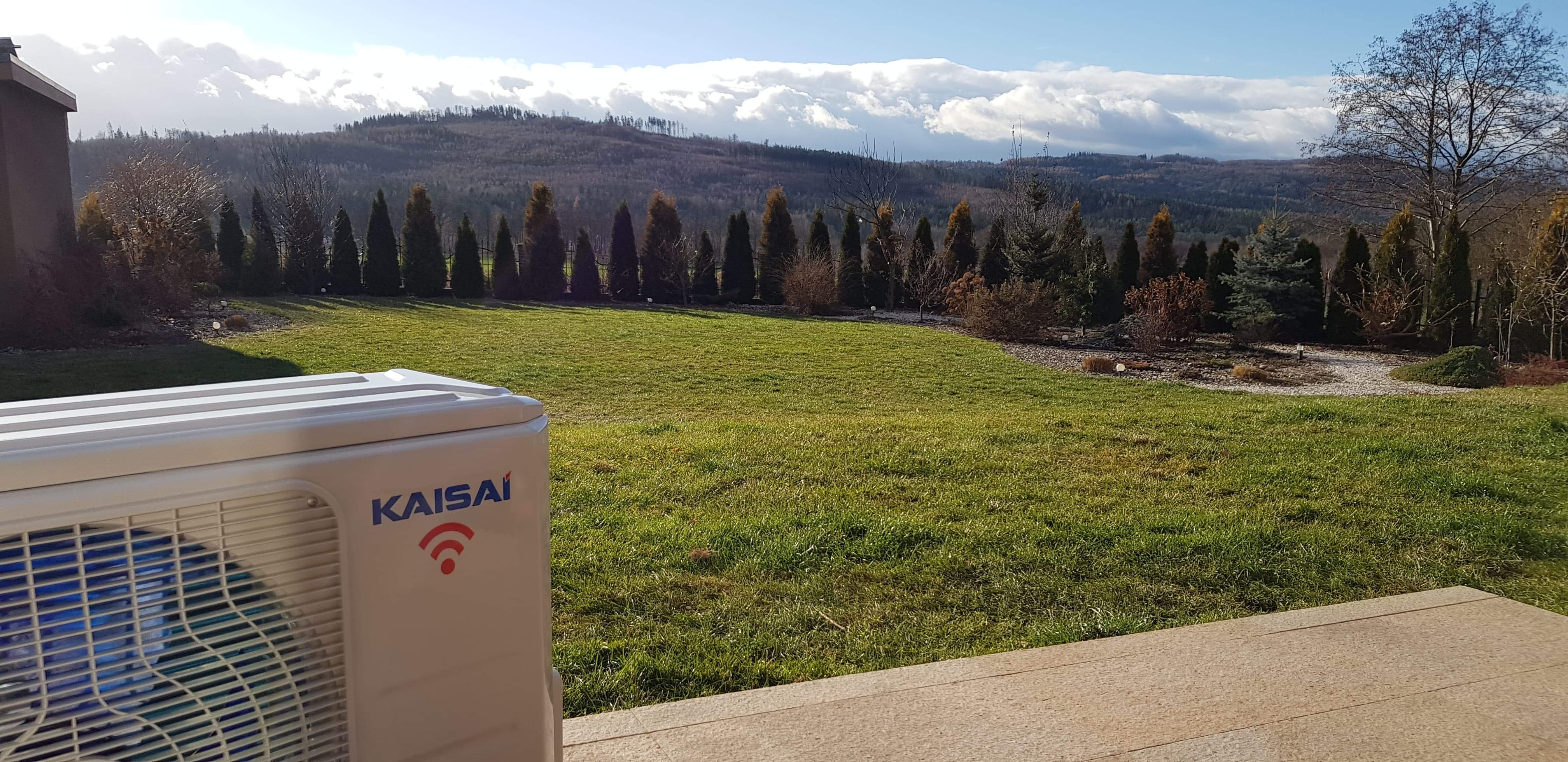 Zewnętrzna jednostka klimatyzacji marki Kaisai na tle zadbanego trawnika, ozdobnych drzew i górskiego krajobrazu pod błękitnym niebem.