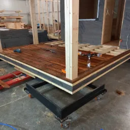 Wnętrze w trakcie budowy: drewniana konstrukcja domu modułowego na metalowej platformie z kółkami, widoczne narzędzia i butelka. Podłoga wyłożona drewnianymi deskami w odcieniach brązu.