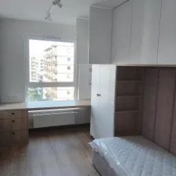 Pokój z zabudową meblową: białe szafy wiszące i stojące, biurko pod oknem z widokiem na budynki mieszkalne, łóżko z zagłówkiem i materacem w folii ochronnej.