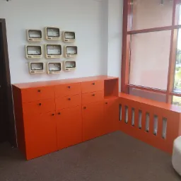 Pomarańczowa szafka z szufladami i szafką obok okna z zabudową grzejnika w tym samym kolorze. Na ścianie dekoracja z obudów starych telewizorów.