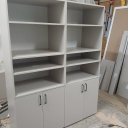 Drewutnia Mazurska Marcin Barczewski - Biała, modułowa szafa z półkami i szafkami, czarne uchwyty, w warsztacie stolarskim, narzędzia na podłodze, surowy montaż, proste linie, minimalistyczny design.