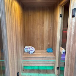 Drewutnia Mazurska Marcin Barczewski - Drewniana sauna z podświetleniem LED, ręcznikami na ławce i oświetleniem sufitowym. Widok z bliska na detale wykończenia i nowoczesny design.