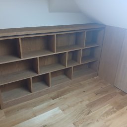 Drewutnia Mazurska Marcin Barczewski - Drewniany regał na wymiar, wkomponowany w skos poddasza, z widoczną strukturą drewna i dopasowaną kolorystyką do podłogi. Widoczne gniazdko elektryczne.