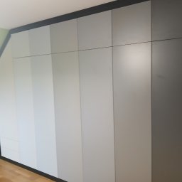 Drewutnia Mazurska Marcin Barczewski - Szafa wnękowa na poddaszu ze skośnym sufitem, fronty w odcieniach szarości i czerni, minimalistyczny design, drewniana podłoga, jasne wnętrze.