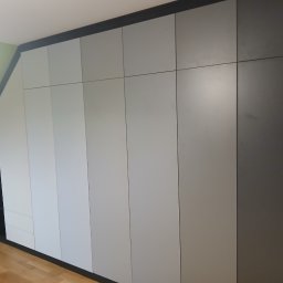 Drewutnia Mazurska Marcin Barczewski - Szara szafa wnękowa z czarnymi akcentami, dopasowana do skosu poddasza, z widocznymi półkami po lewej stronie i drewnianą podłogą.