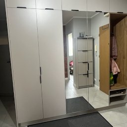 Drewutnia Mazurska Marcin Barczewski - Jasna szafa wnękowa z lustrem i czarnymi uchwytami, zintegrowana z wieszakiem na ubrania. Widoczny fragment przedpokoju z szarą wycieraczką.