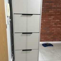 Drewutnia Mazurska Marcin Barczewski - Wysoka, wąska szafka na buty z czterema szufladami w odcieniach szarości, z czarnymi uchwytami, na tle ściany z czerwonej cegły i jasnej podłogi.