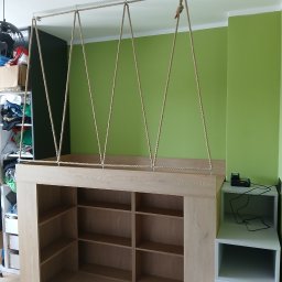 Drewutnia Mazurska Marcin Barczewski - Kreatywna konstrukcja z jasnego drewna z półkami, zawieszona na linach przymocowanych do sufitu, na tle zielonej ściany. Nietypowy mebel w nowoczesnym wnętrzu.