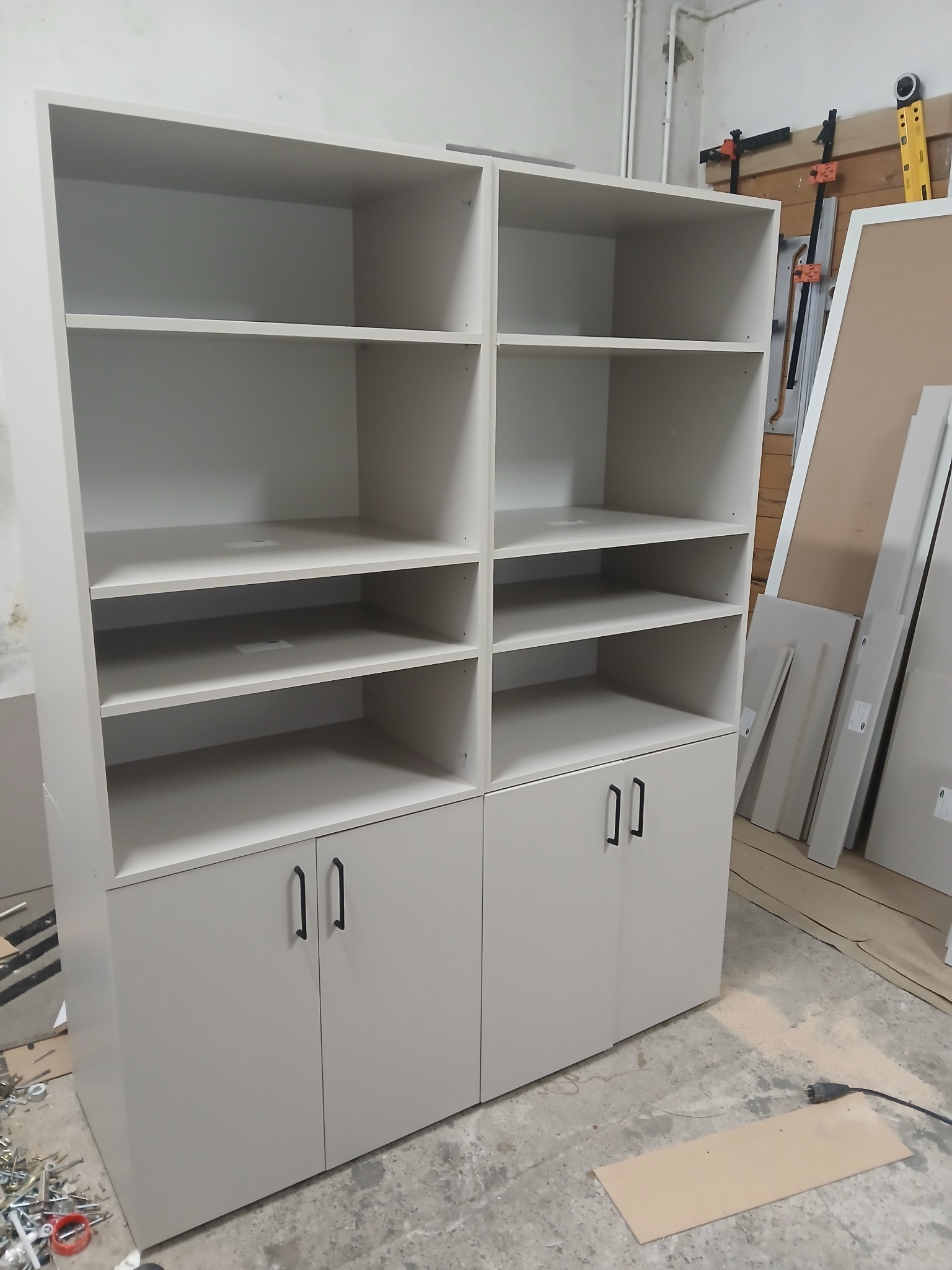 Biała, modułowa szafa z półkami i szafkami, czarne uchwyty, w warsztacie stolarskim, narzędzia na podłodze, surowy montaż, proste linie, minimalistyczny design.